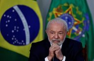 Presidente Lula com barba e terno escuro, mãos unidas, sentado à frente de bandeiras do Brasil.