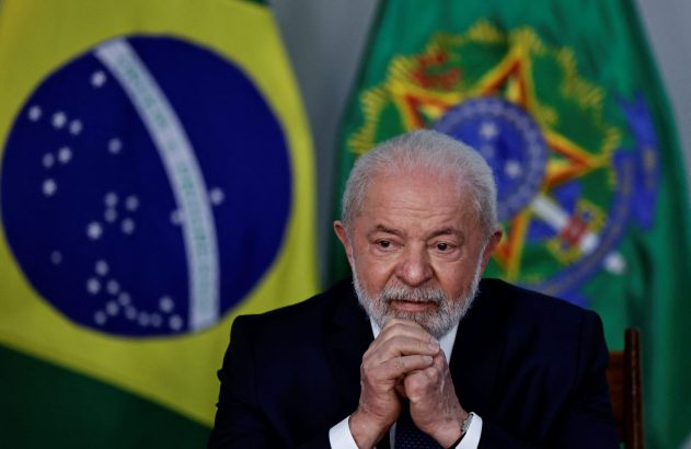 Presidente Lula com barba e terno escuro, mãos unidas, sentado à frente de bandeiras do Brasil.