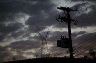 Poste com transformador e torre de transmissão em silhueta sob céu escuro e nublado.