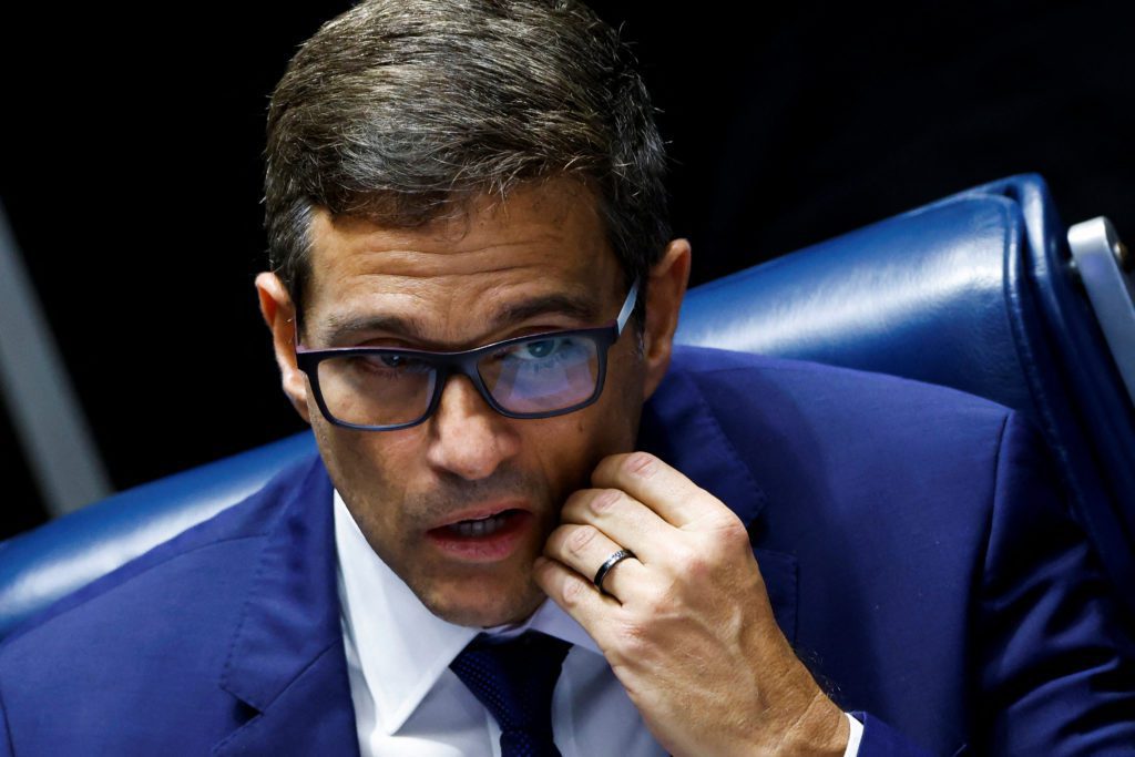 Homem de terno azul e óculos, com a mão no queixo e boca entreaberta, em pose reflexiva.