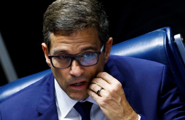 Homem de terno azul e óculos, com a mão no queixo e boca entreaberta, em pose reflexiva.