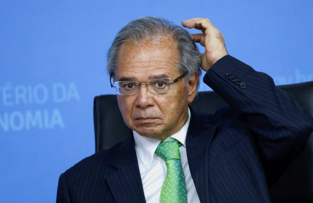 Homem de terno, óculos e gravata verde coça a cabeça com semblante pensativo.