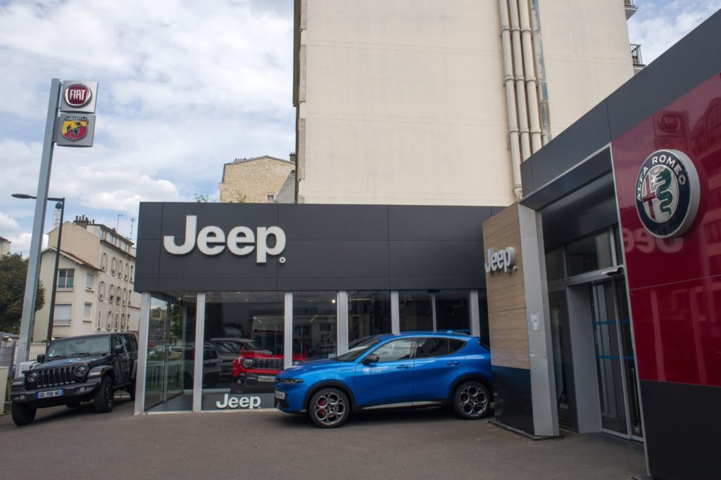 Concessionária de carros com marcas Jeep, Alfa Romeo, Fiat e Abarth. Um Jeep preto e um SUV azul estacionados.