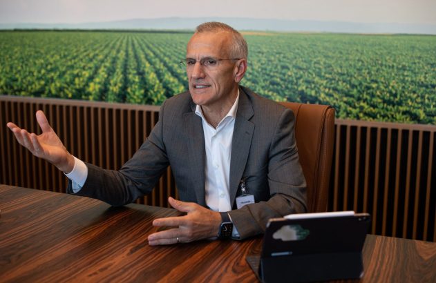 Homem de terno cinza sentado à mesa de madeira escura, gesticula. Ao fundo, grande imagem de um vasto campo agrícola verde.