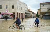Dois homens pedalam bicicletas em rua de cidade alagada por água turva. Um carrossel está parcialmente imerso na água à esquerda.