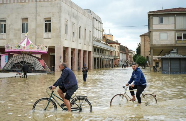 Dois homens pedalam bicicletas em rua de cidade alagada por água turva. Um carrossel está parcialmente imerso na água à esquerda.