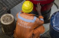 Homem em macacão ONGC laranja e capacete amarelo agachado, tocando equipamento vermelho. No uniforme: 'Safe Way is the Right Way'.