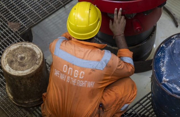 Homem em macacão ONGC laranja e capacete amarelo agachado, tocando equipamento vermelho. No uniforme: 'Safe Way is the Right Way'.