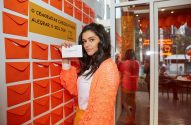 Mulher de cardigan laranja mostra cartão com mensagem em parede de envelopes e letreiro Cenoradas. Loja ao fundo.