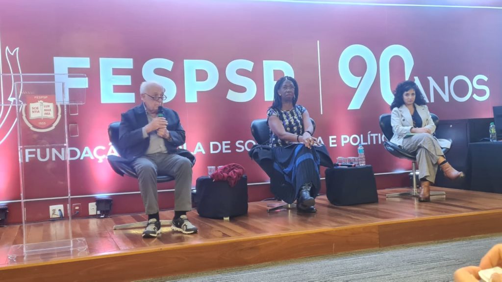 Três palestrantes sentados no palco em evento FESPSP 90 anos, homem com microfone.