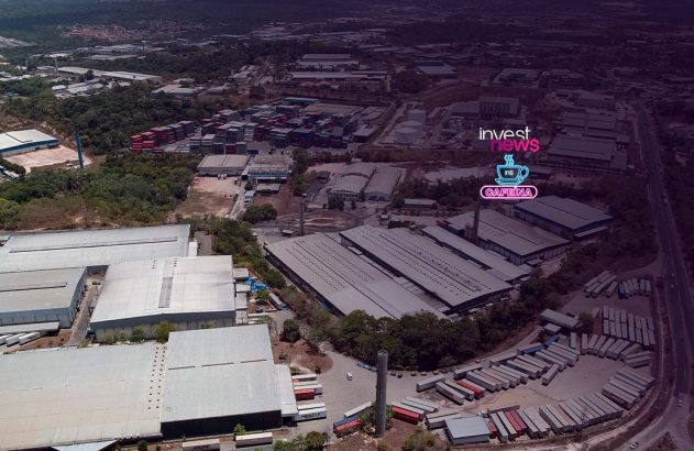 Vista aérea de complexo industrial com galpões. Logo "Invest News" com xícara de café sobreposto digitalmente.
