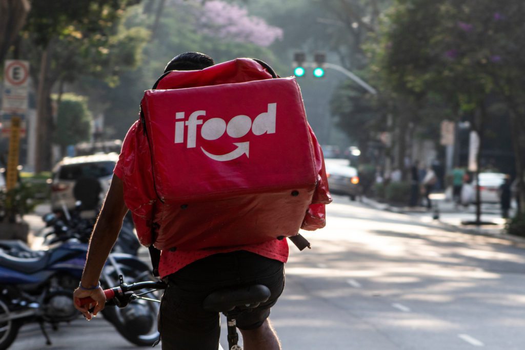 Ciclista entregador do iFood, de costas, pedalando em uma rua urbana ensolarada.