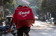 Ciclista entregador do iFood, de costas, pedalando em uma rua urbana ensolarada.