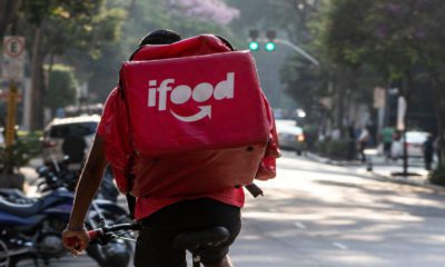 Ciclista entregador do iFood, de costas, pedalando em uma rua urbana ensolarada.