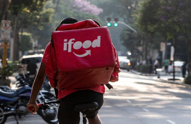 Ciclista entregador do iFood, de costas, pedalando em uma rua urbana ensolarada.