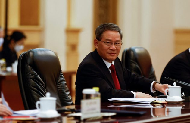 Homem asiático de óculos e terno em mesa de reunião, olhando para a câmera com sorriso.