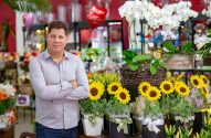 Homem sorridente de braços cruzados em floricultura, rodeado por orquídeas brancas e girassóis amarelos.