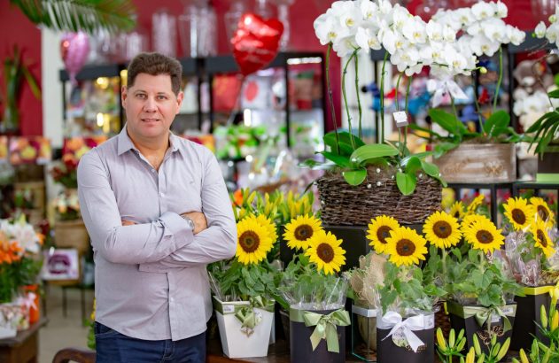 Homem sorridente de braços cruzados em floricultura, rodeado por orquídeas brancas e girassóis amarelos.