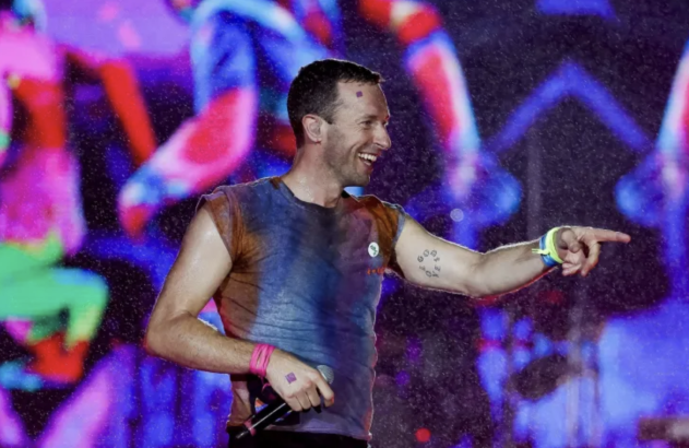 Homem sorrindo, apontando e com microfone, em palco. Gotas de água voam; luzes vibrantes iluminam o show.