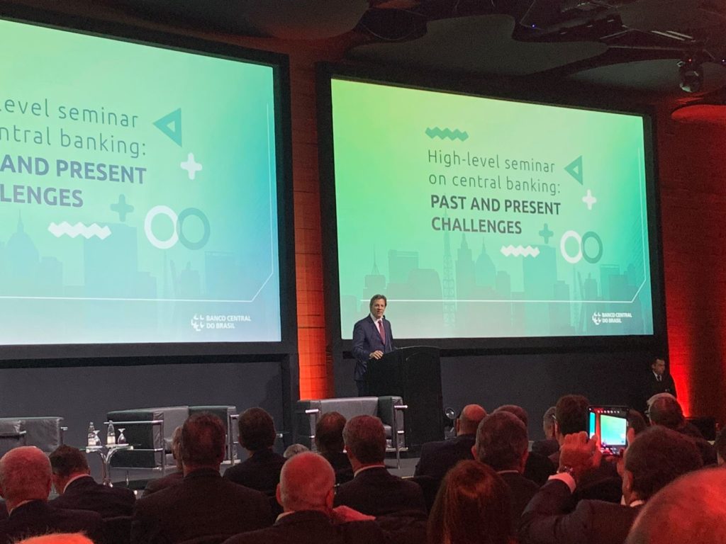 Homem discursando em seminário do Banco Central do Brasil. Telas exibem: "High-level seminar on central banking: PAST AND PRESENT CHALLENGES".