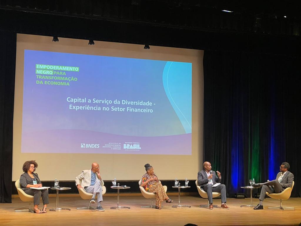 Cinco painelistas diversos debatem empoderamento negro e diversidade financeira em palco. Tema exibido em tela grande.