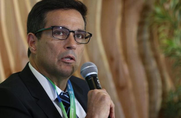 Homem de terno, óculos e crachá falando ao microfone.