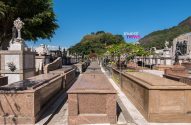 Cemitério com múltiplas sepulturas em fileiras, algumas com esculturas, sob céu azul e montanhas arborizadas.