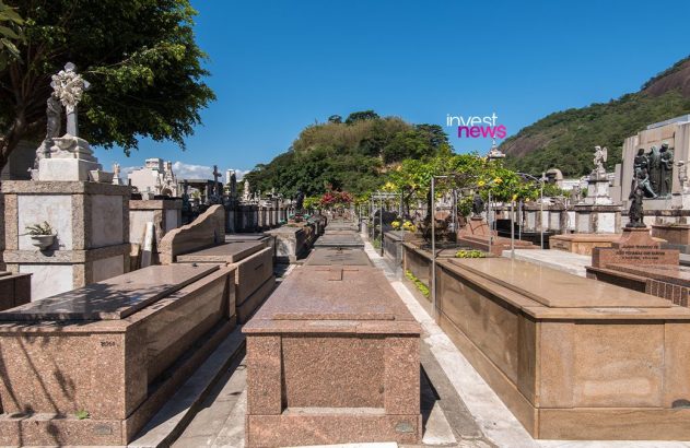 Cemitério com múltiplas sepulturas em fileiras, algumas com esculturas, sob céu azul e montanhas arborizadas.