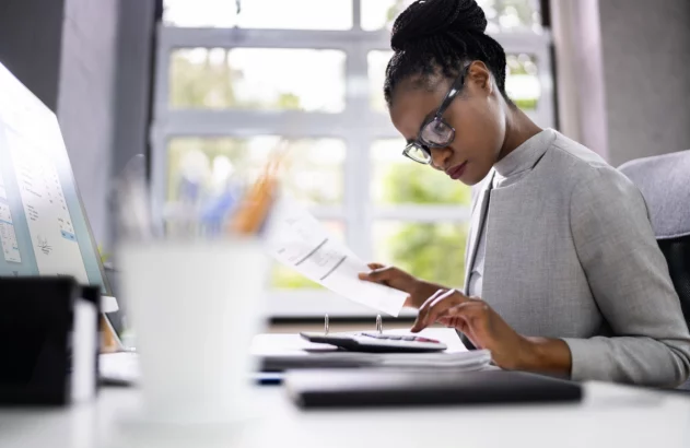 Mulher negra de blazer cinza em frente a um computador, fazendo contas em uma calculadora com um documento em mãos