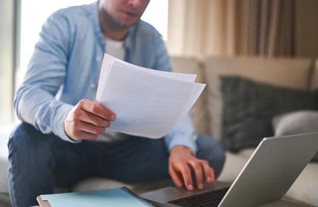 Homem teclando em um notebook com uma mão, enquanto a outra segura algumas folhas de documentos