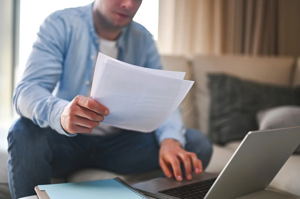 Homem teclando em um notebook com uma mão, enquanto a outra segura algumas folhas de documentos