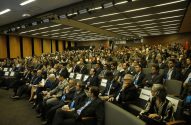Amplo auditório lotado com centenas de participantes sentados, assistindo a uma conferência.