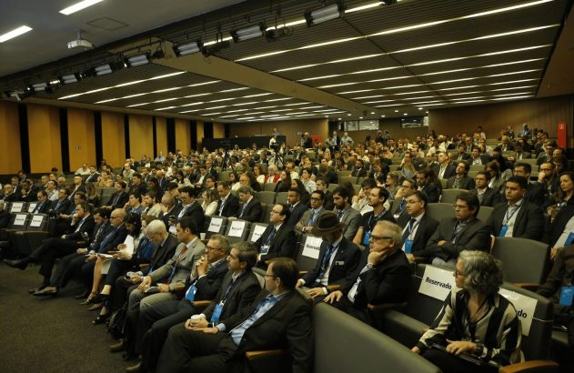 Amplo auditório lotado com centenas de participantes sentados, assistindo a uma conferência.