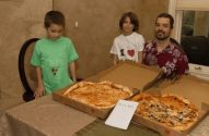 Duas crianças e um homem olham pizzas em três caixas sobre uma mesa.