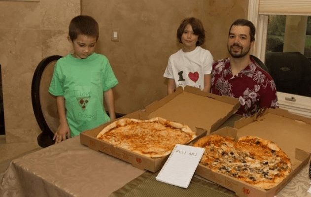 Duas crianças e um homem olham pizzas em três caixas sobre uma mesa.