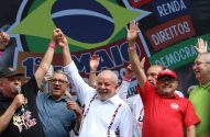 Lula sorri, levanta mãos com outro líder em comício. Demais em palco, banner "1º MAIO" ao fundo.