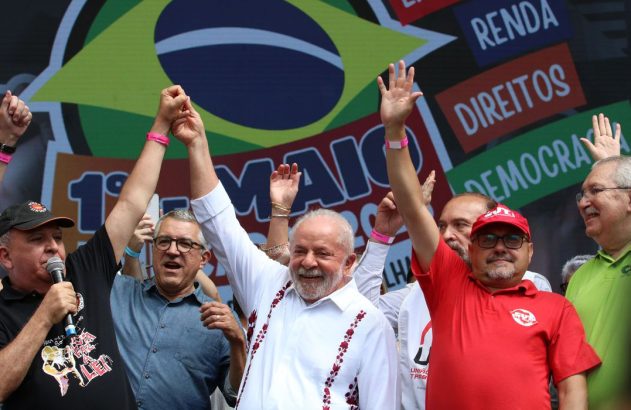 Lula sorri, levanta mãos com outro líder em comício. Demais em palco, banner "1º MAIO" ao fundo.