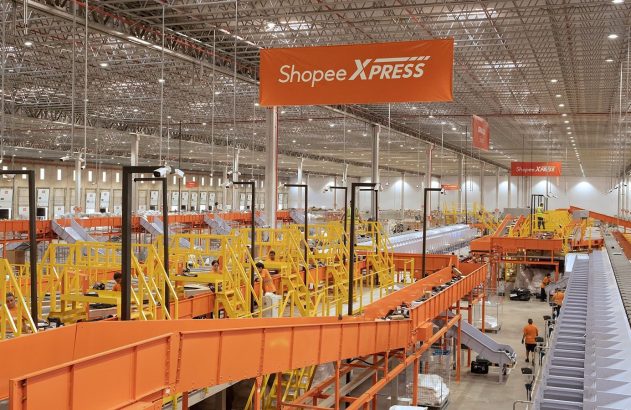 Centro de logística Shopee XPRESS com esteiras transportando e classificando pacotes. Funcionários operam o sistema.