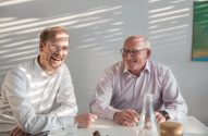 Dois homens sorrindo, um jovem e um mais velho, com óculos, sentados à mesa.