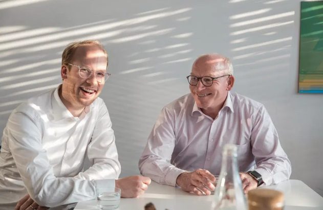 Dois homens sorrindo, um jovem e um mais velho, com óculos, sentados à mesa.