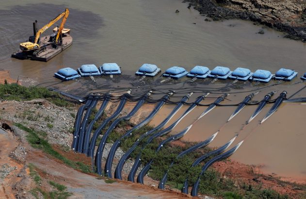 Operação de dragagem: escavadeira em água turva bombeia por tubos para sacos de filtragem grandes na margem barrenta.