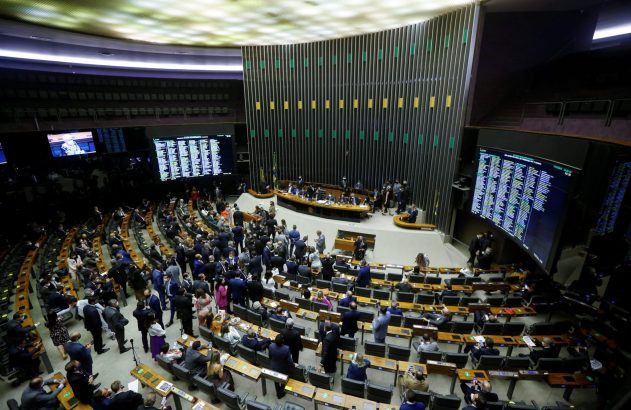 Plenário de assembleia legislativa durante sessão. Pessoas sentadas e em pé, telas com dados visíveis.