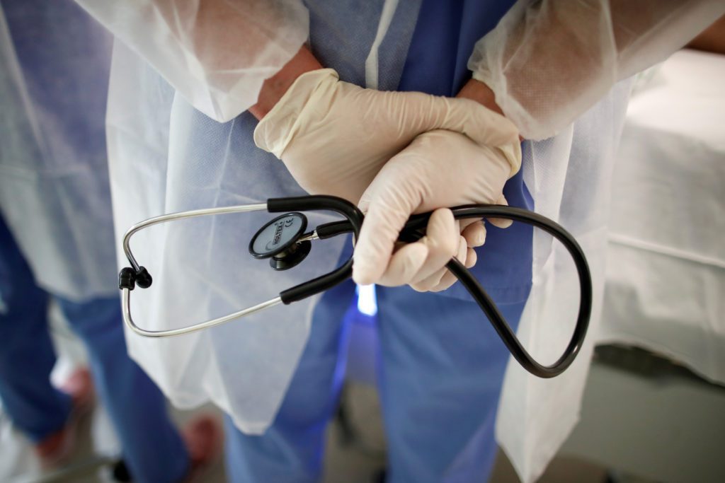 Profissional de saúde em avental e luvas, segurando um estetoscópio com as mãos atrás das costas.
