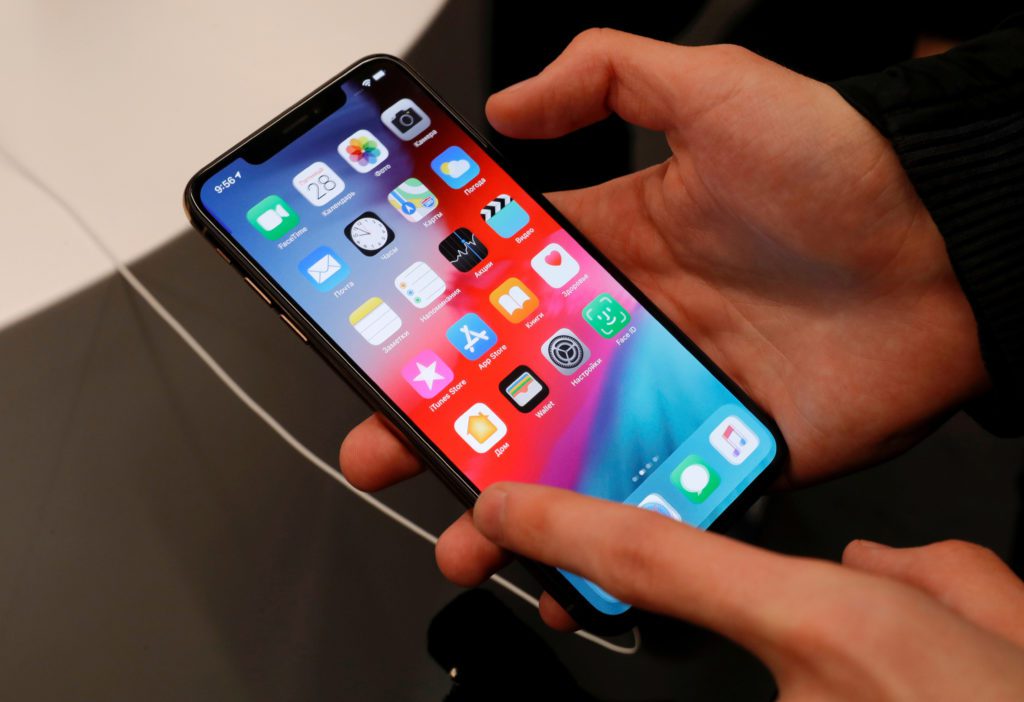 Mãos segurando e usando um iPhone com a tela ligada exibindo ícones de aplicativos.