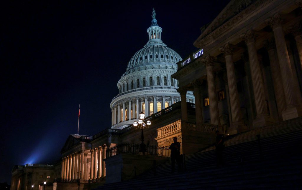 Capitólio dos EUA à noite, cúpula e fachadas iluminadas. Há figuras em escadarias amplas e uma bandeira americana.