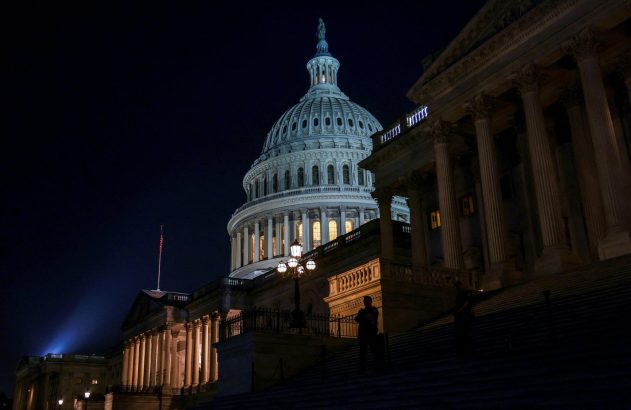 Capitólio dos EUA à noite, cúpula e fachadas iluminadas. Há figuras em escadarias amplas e uma bandeira americana.