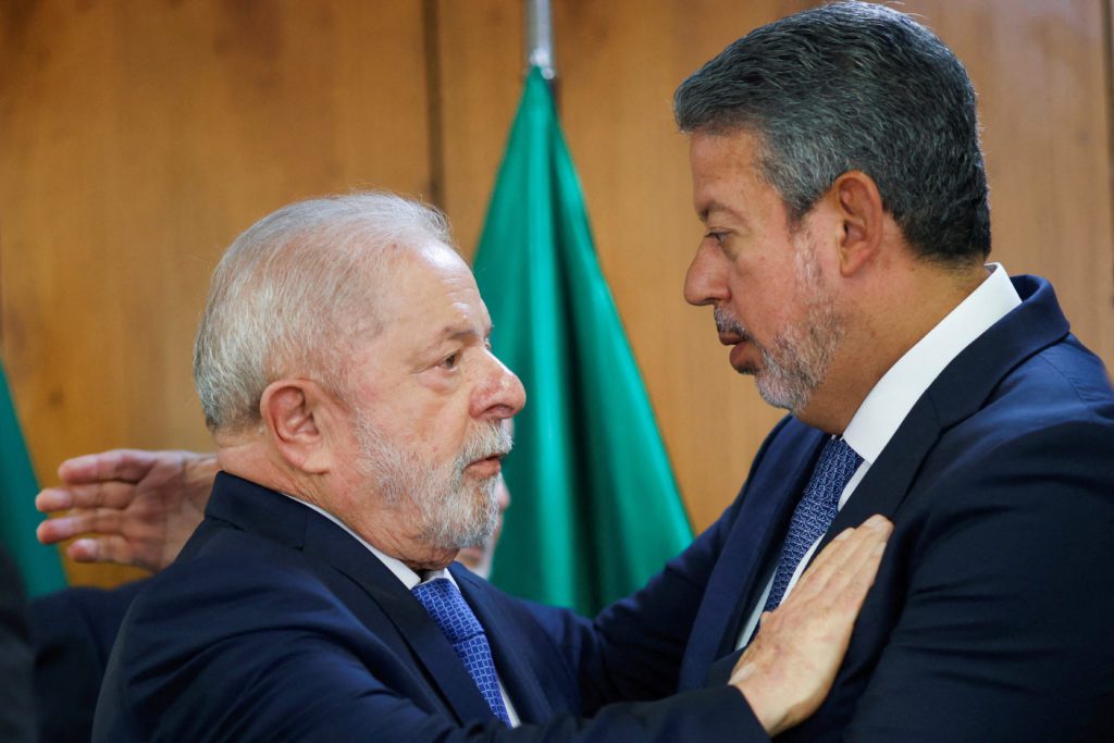 Dois homens em ternos escuros, de perfil, se olham de perto com as mãos nos ombros. Bandeira verde.