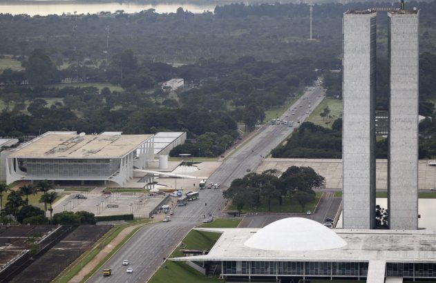 Vista aérea de Brasília: edifícios governamentais modernistas (torres, cúpula) e avenida com veículos.