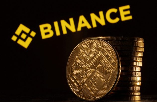 Moeda de criptomoeda dourada em destaque com uma pilha de moedas e o logo da Binance iluminado ao fundo.
