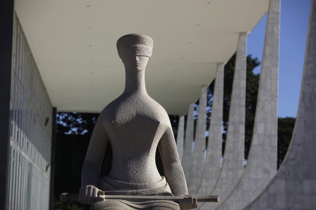 Estátua de pedra da Justiça, vendada e sentada, em frente a um edifício modernista com colunas sob o céu azul.